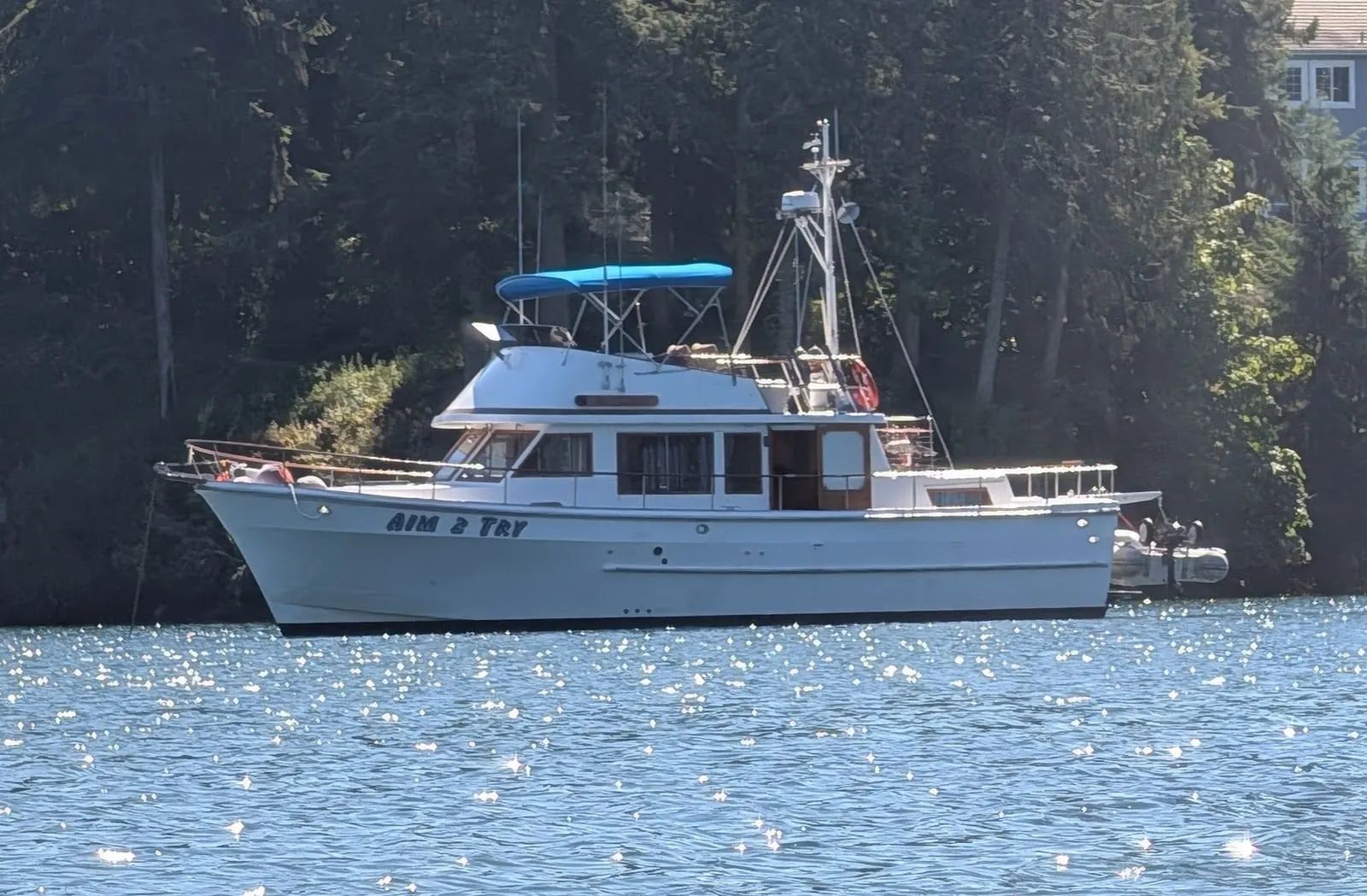 Aim 2 Try, a white 41 ft CHB trawler with a tall house, a flybridge under a blue bimini and a dinghy on the stern, lying to a mooring line in front of a tree-lined shore.