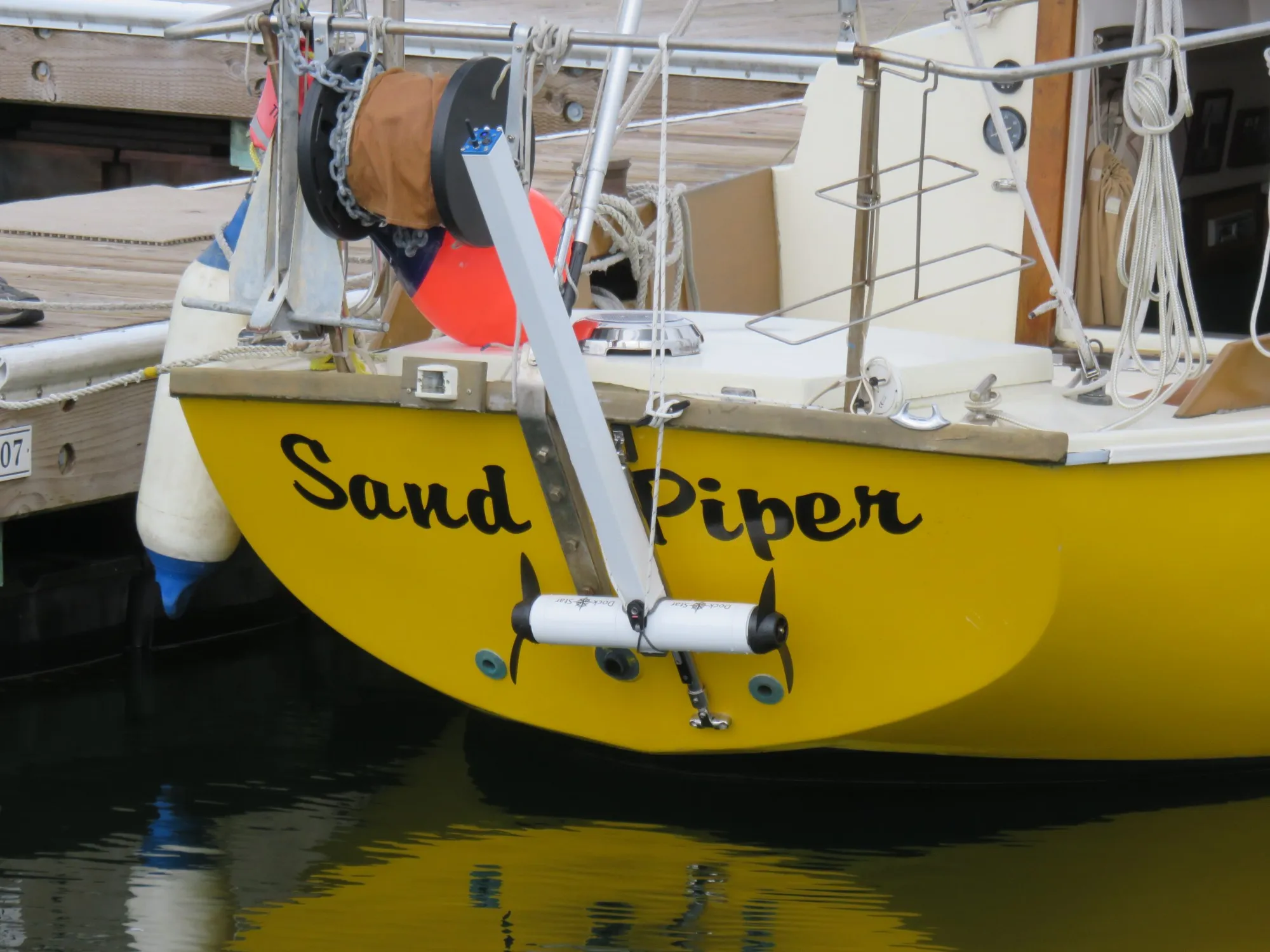 The yellow stern of a sailboat at a dock, with a DockStar on a track on the transom, tilted up so its white pod and two black propellers hang just above the water.